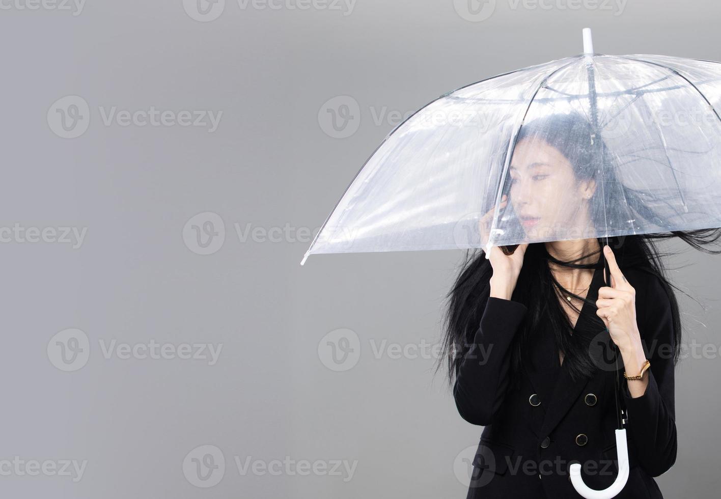 femme transgenre asiatique aux longs cheveux raides noirs, coup de vent jeté dans l'air. femme tenir le téléphone et le parapluie contre la tempête de vent, se sentir mode sensuelle sexy, espace de copie isolé sur fond gris photo