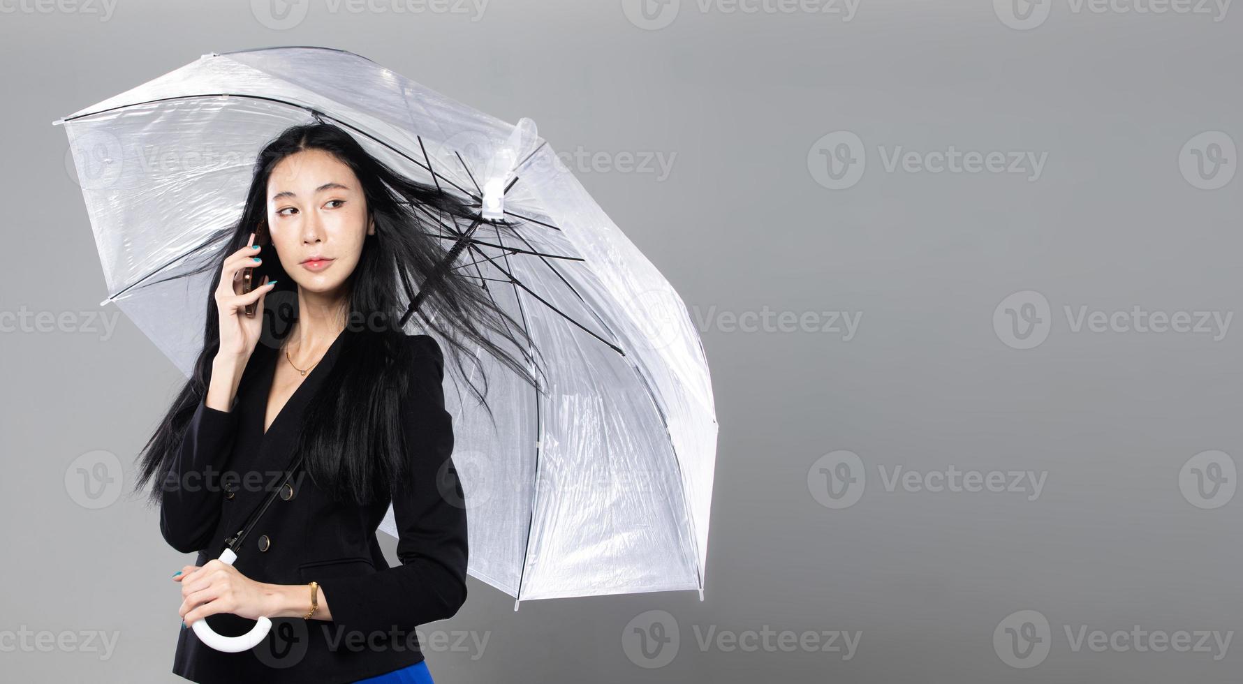 femme transgenre asiatique aux longs cheveux raides noirs, coup de vent jeté dans l'air. femme tenir le téléphone et le parapluie contre la tempête de vent, se sentir mode sensuelle sexy, espace de copie isolé sur fond gris photo