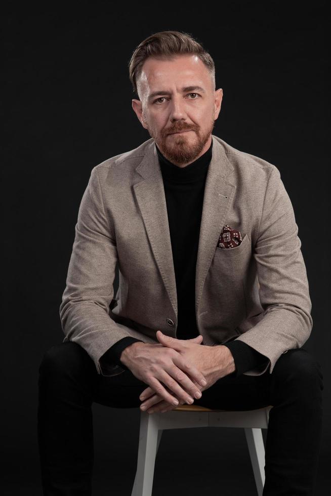 portrait d'un homme d'affaires adulte portant un costume à la mode et assis dans un studio moderne sur une chaise élégante sur fond noir photo