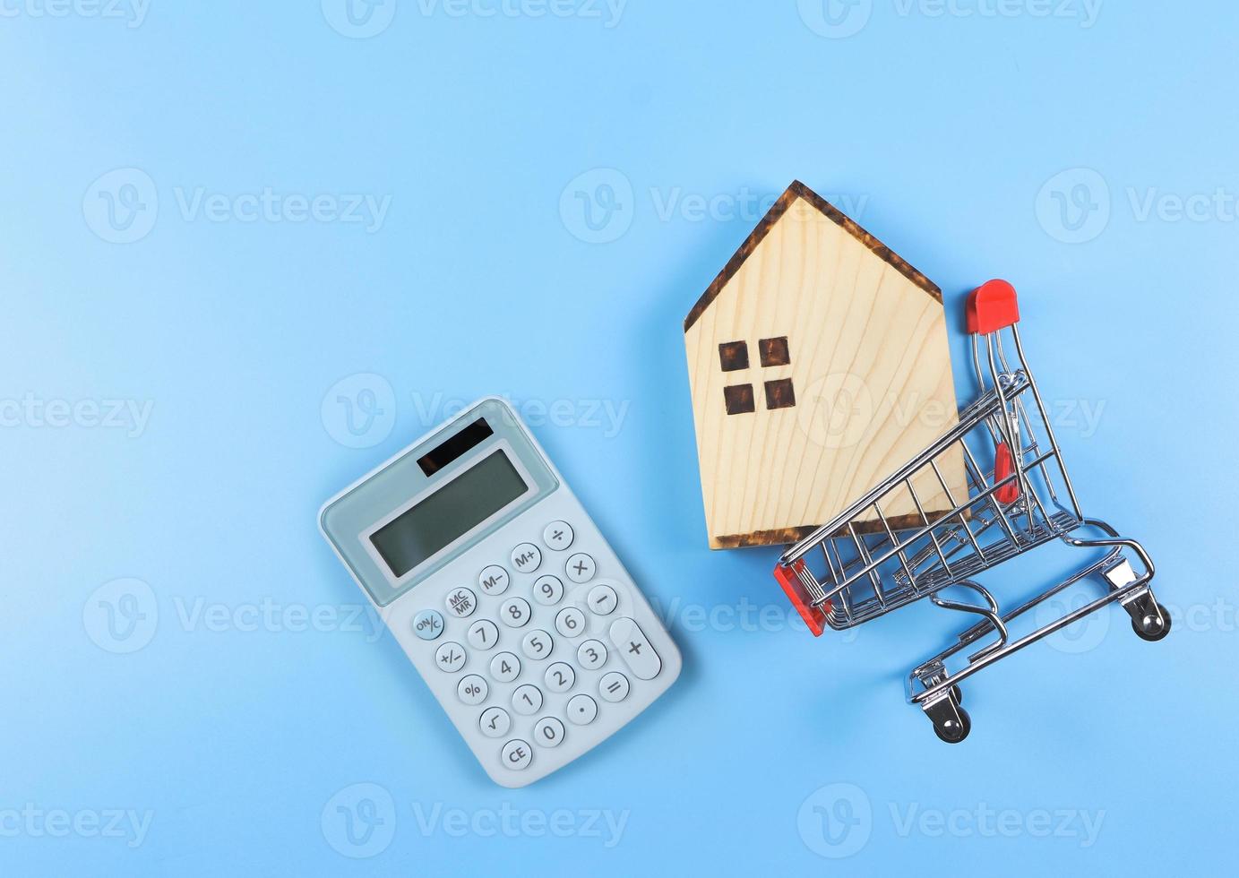mise en page à plat du modèle de maison en bois dans le caddie et calculatrice bleue sur fond bleu avec espace de copie, concept d'achat de maison. photo