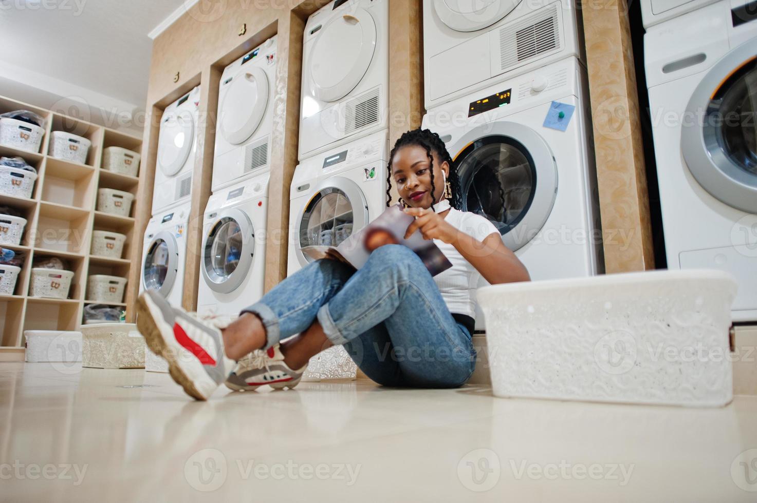 joyeuse femme afro-américaine assise avec des écouteurs et lisant un magazine près de la machine à laver dans la laverie en libre-service. photo