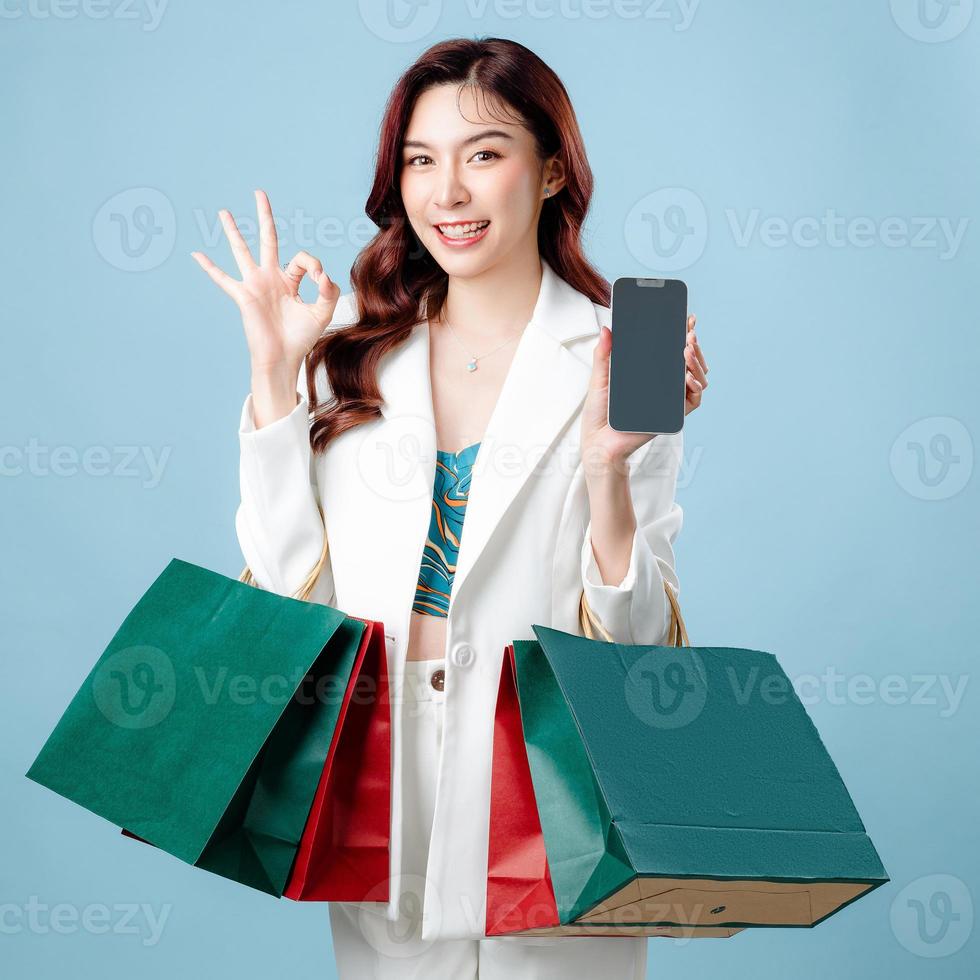 la moitié du corps d'une belle femme d'affaires asiatique portant un costume blanc montre un signe ok tenant une application de téléphonie mobile et un sac à provisions sur fond bleu isolé. concept de smartphone à écran blanc. photo
