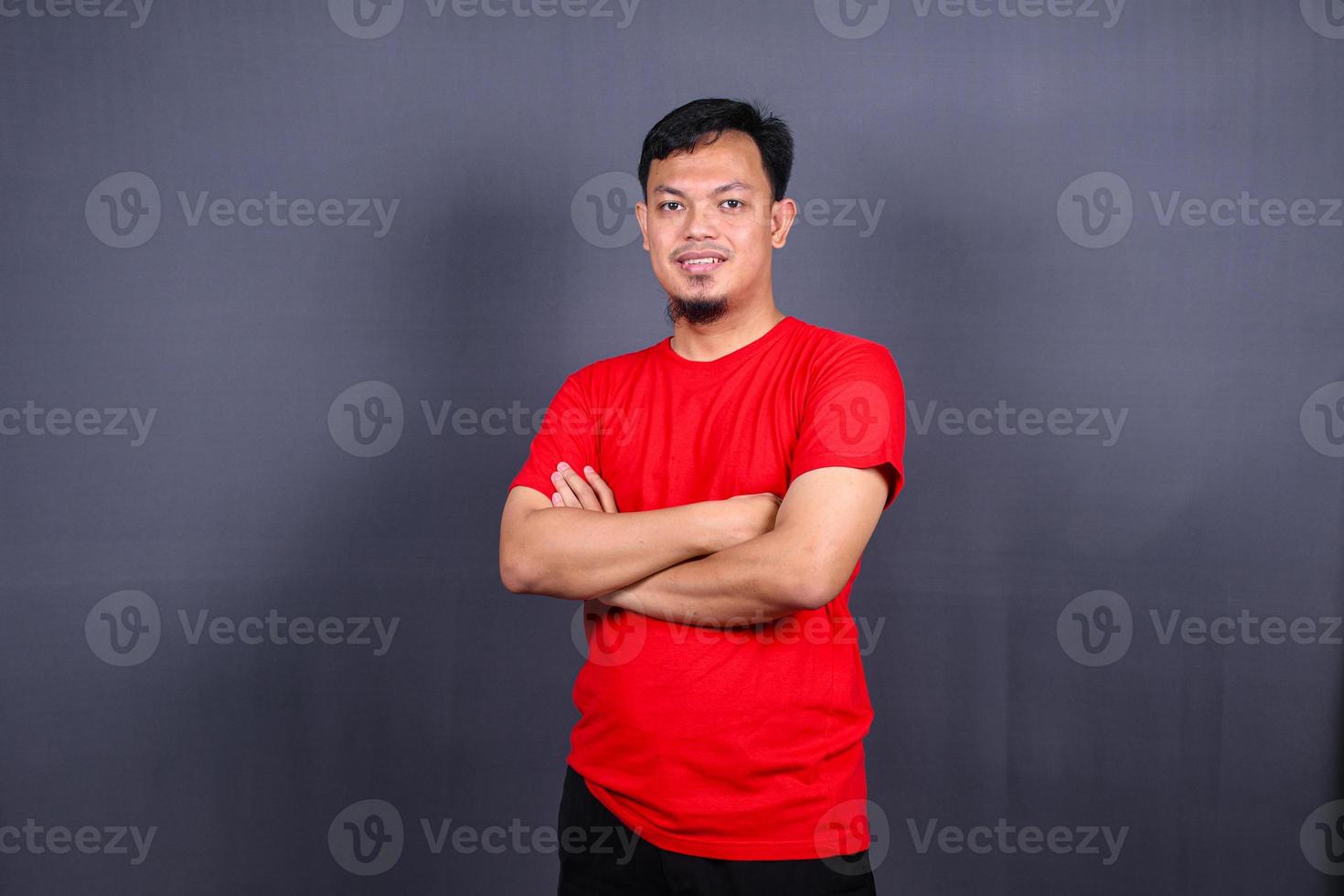 portrait d'un homme asiatique séduisant en t-shirt rouge debout avec les bras croisés isolé sur fond gris photo