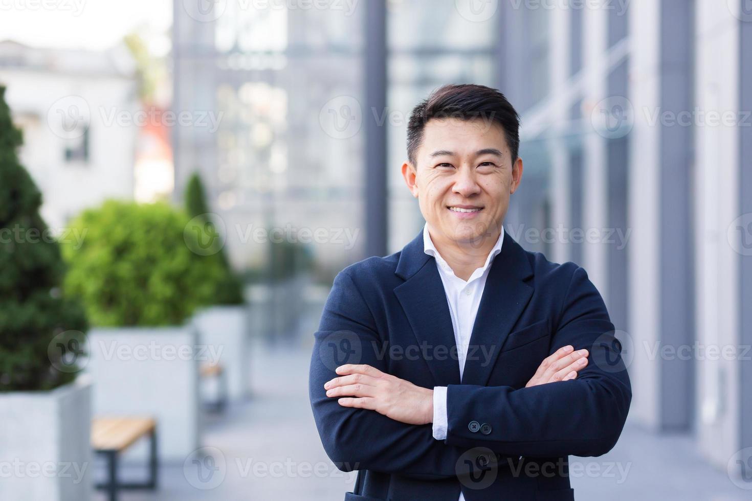 patron asiatique réussi près du bureau regarde la caméra avec des sourires de bras croisés photo