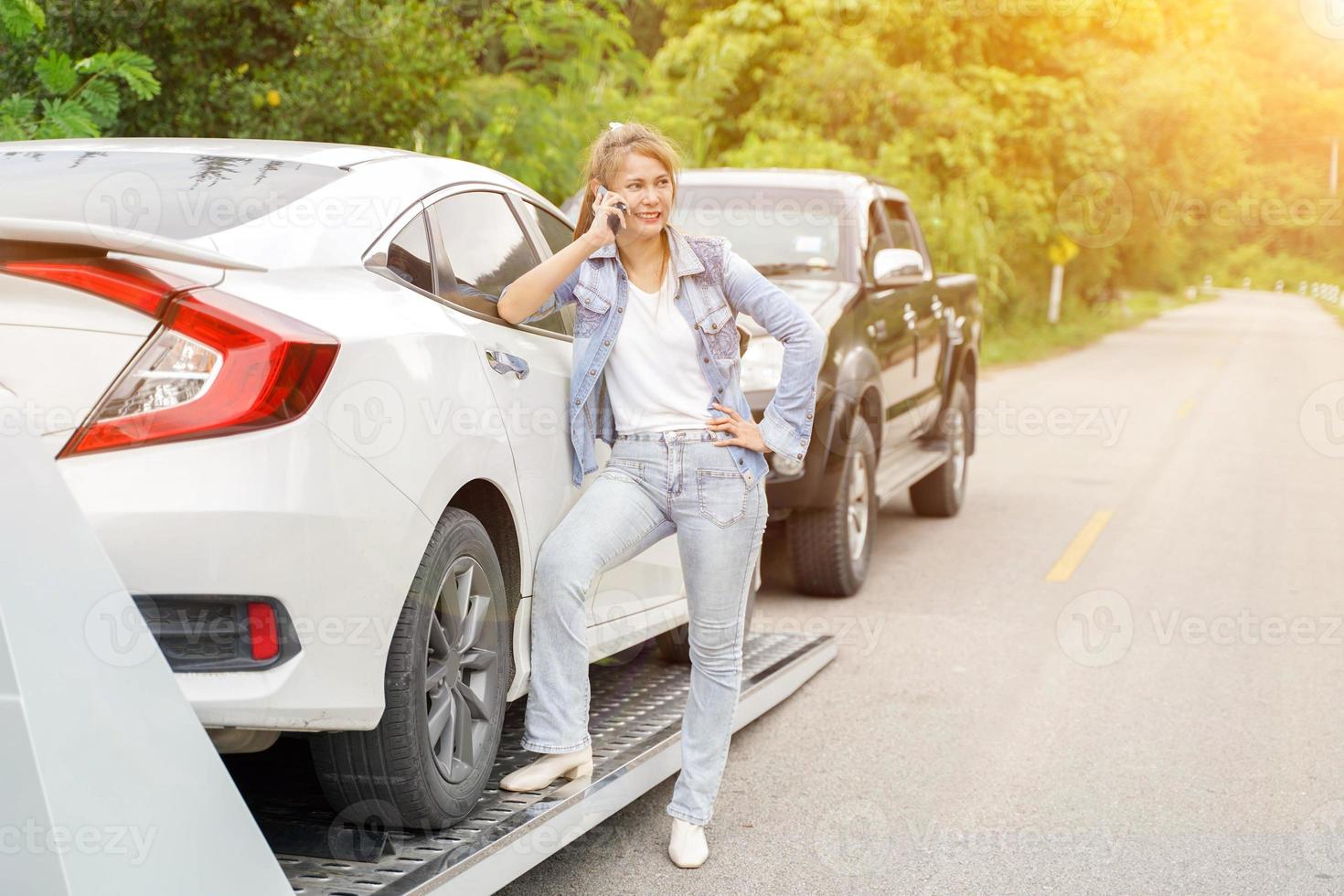 jeune femme faisant le téléphone appelant quelqu'un pendant le service de remorquage l'aidant sur la route de campagne avec la lumière du soleil. concept d'assistance routière. photo