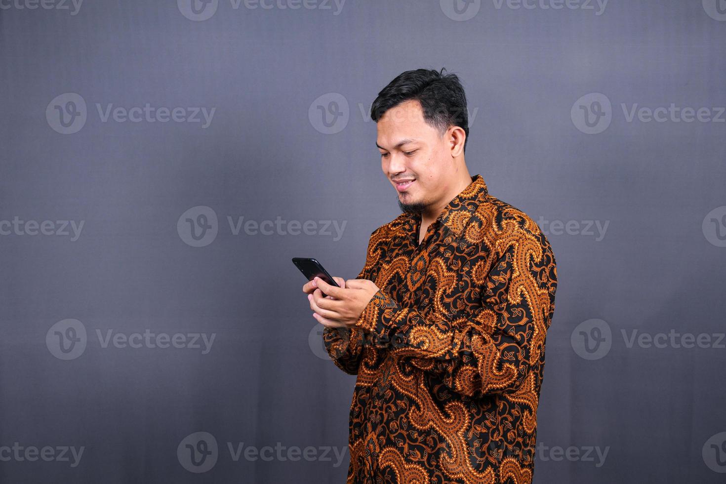 portrait d'un jeune homme asiatique portant du batik à l'aide d'un téléphone portable sur fond gris photo