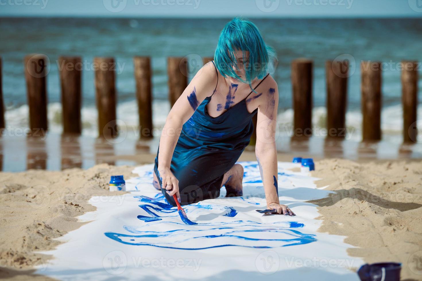 Artiste de performance artistique femme aux cheveux bleus enduite de peintures à la gouache sur une grande toile sur la plage photo