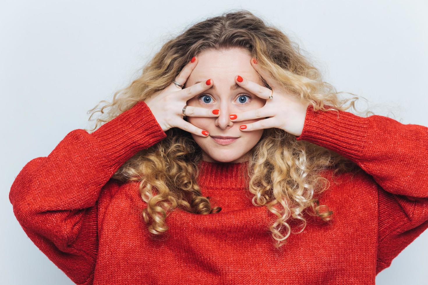 portrait horizontal d'une mystérieuse femme aux yeux bleus avec des cheveux clairs bouclés, couvre le visage avec les mains, vêtue d'un pull rouge ample, isolée sur fond blanc. concept de surprise et de personnes photo