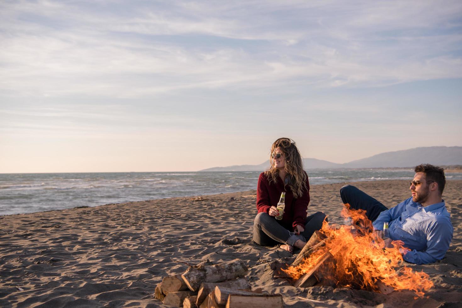 jeune couple assis sur la plage à côté d'un feu de camp buvant de la bière photo