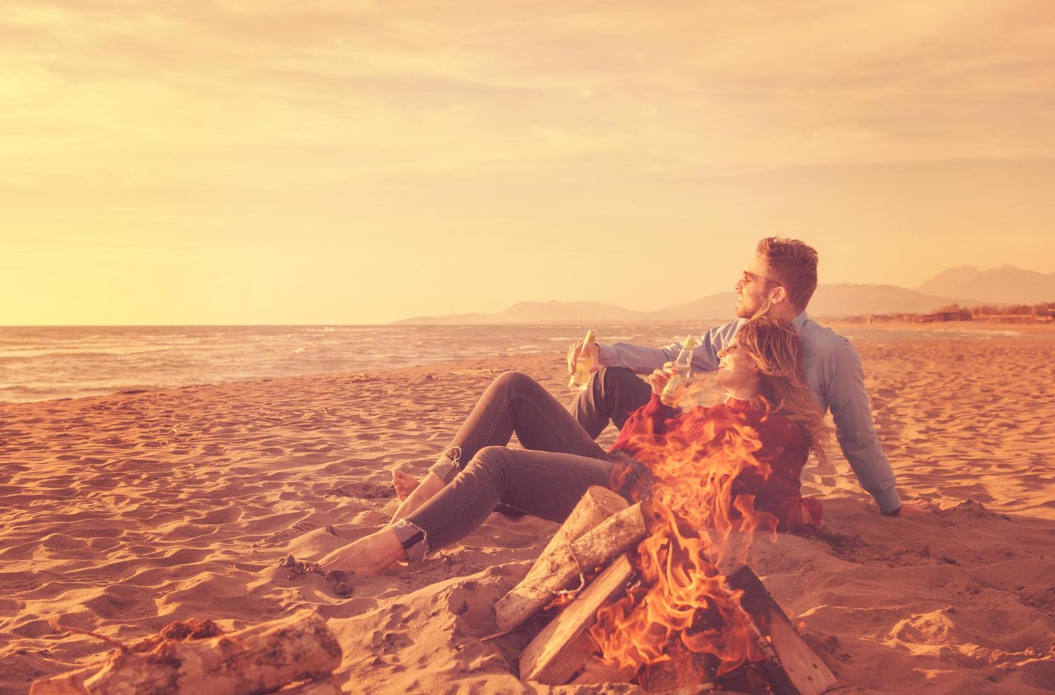 jeune couple assis sur la plage à côté d'un feu de camp buvant de la bière photo