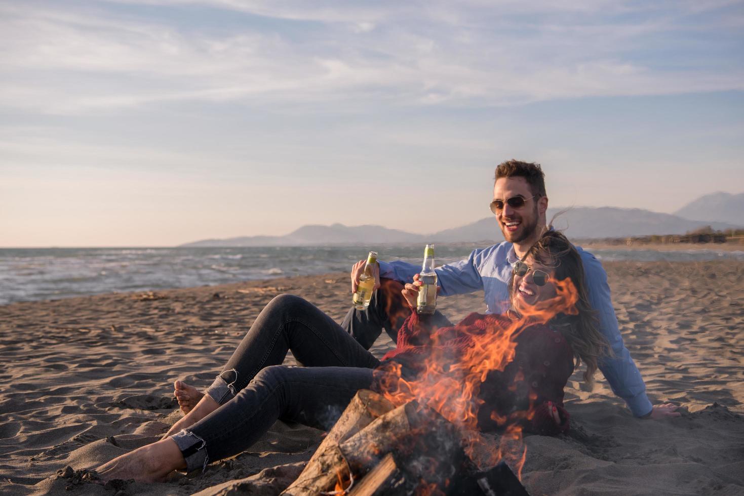 jeune couple assis sur la plage à côté d'un feu de camp buvant de la bière photo