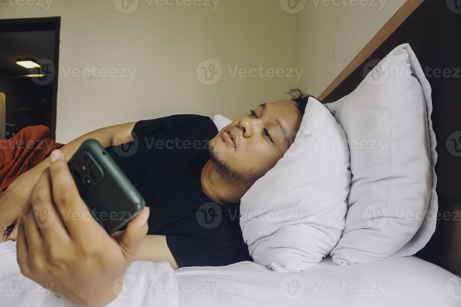 jeune homme fatigué vérifiant son téléphone en position couchée dans son lit photo