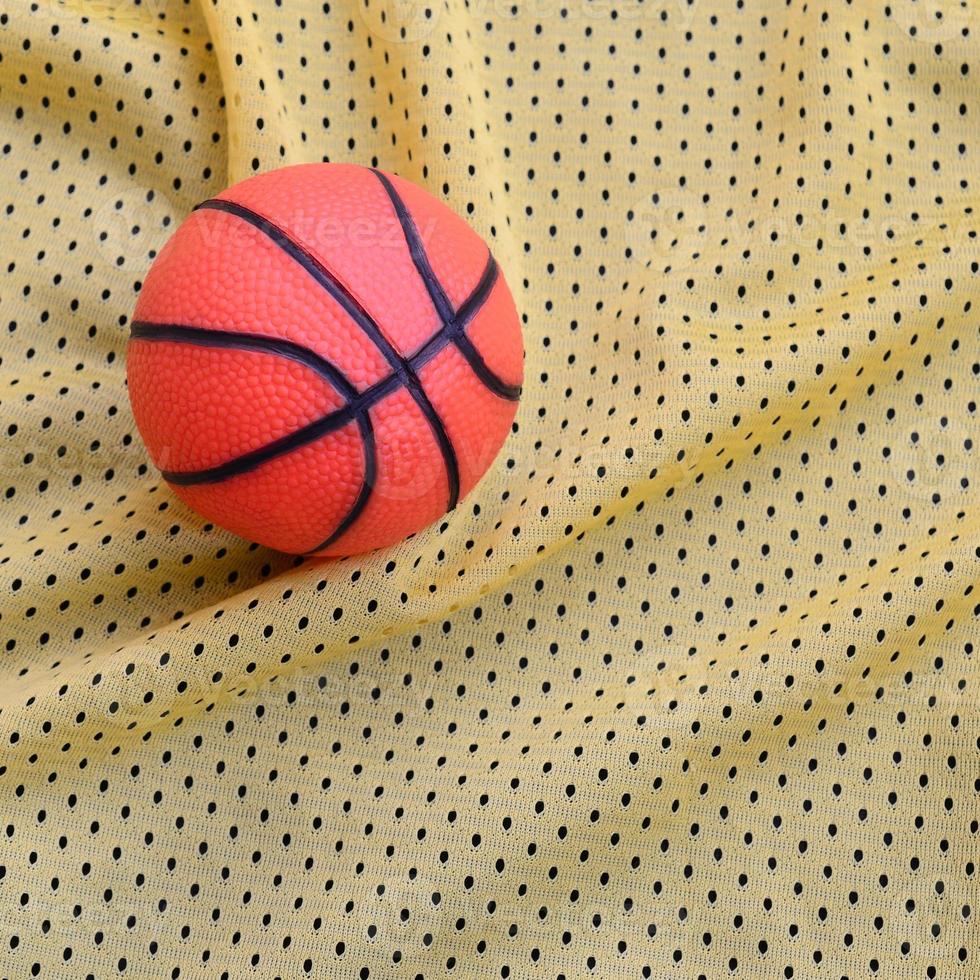 petit basket-ball en caoutchouc orange se trouve sur une texture et un fond de tissu de vêtements de maillot de sport jaune avec de nombreux plis photo