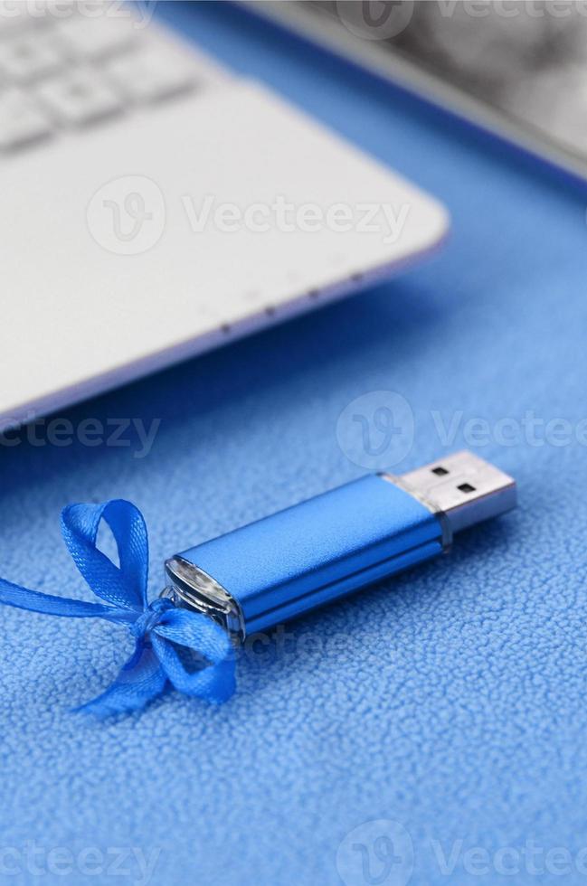 carte mémoire flash usb bleu brillant avec un arc bleu se trouve sur une couverture de tissu polaire bleu clair doux et poilu à côté d'un ordinateur portable blanc et d'un smartphone. conception de cadeau féminin classique pour une carte mémoire photo