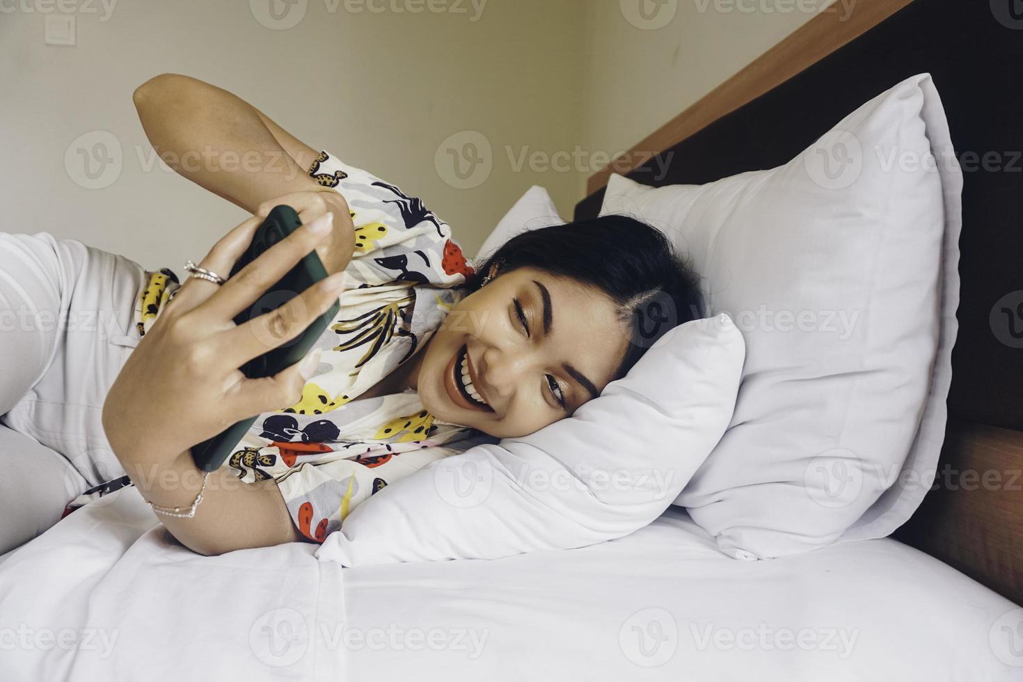 heureuse jeune femme vérifiant son téléphone en position couchée dans son lit photo