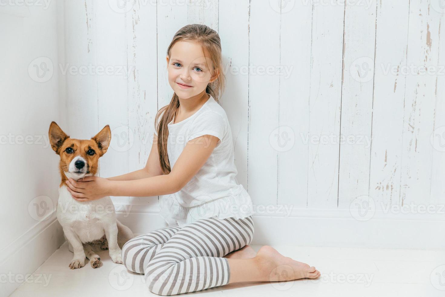 une jolie petite fille joue avec son chien dans une salle blanche, s'assoit par terre, a de bonnes relations, câline son animal de compagnie préféré. petite écolière aime les animaux, passe du temps libre à la maison. enfance photo