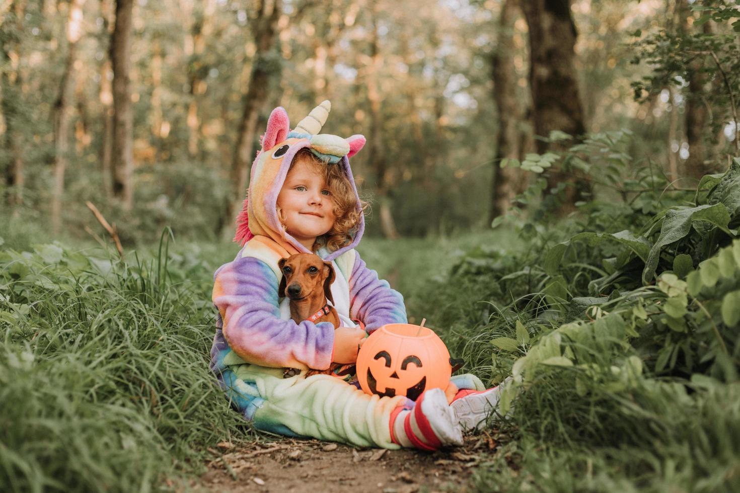 petite fille qui rit et un teckel nain en costumes d'halloween avec un panier de citrouille pour les bonbons à l'extérieur. une fille en costume de kigurumi de licorne arc-en-ciel, un chien en robe avec une jupe ample. vue de dessus photo