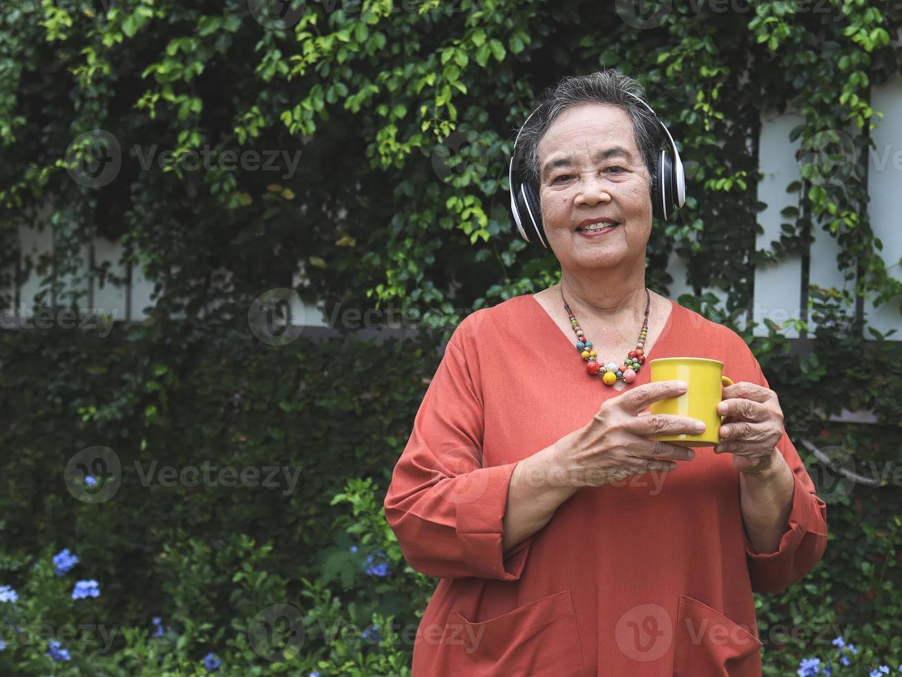 femme asiatique âgée heureuse et en bonne santé tenant une tasse de café jaune debout dans le jardin, écoutant la musique des écouteurs, souriant et regardant la caméra. photo