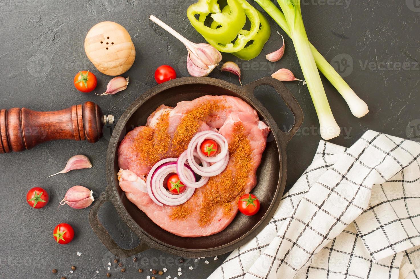 steak cru, entrecôte aux épices pour la cuisson de la viande dans une poêle en fonte avec oignons, ail et tomate. fond noir. vue de dessus. photo
