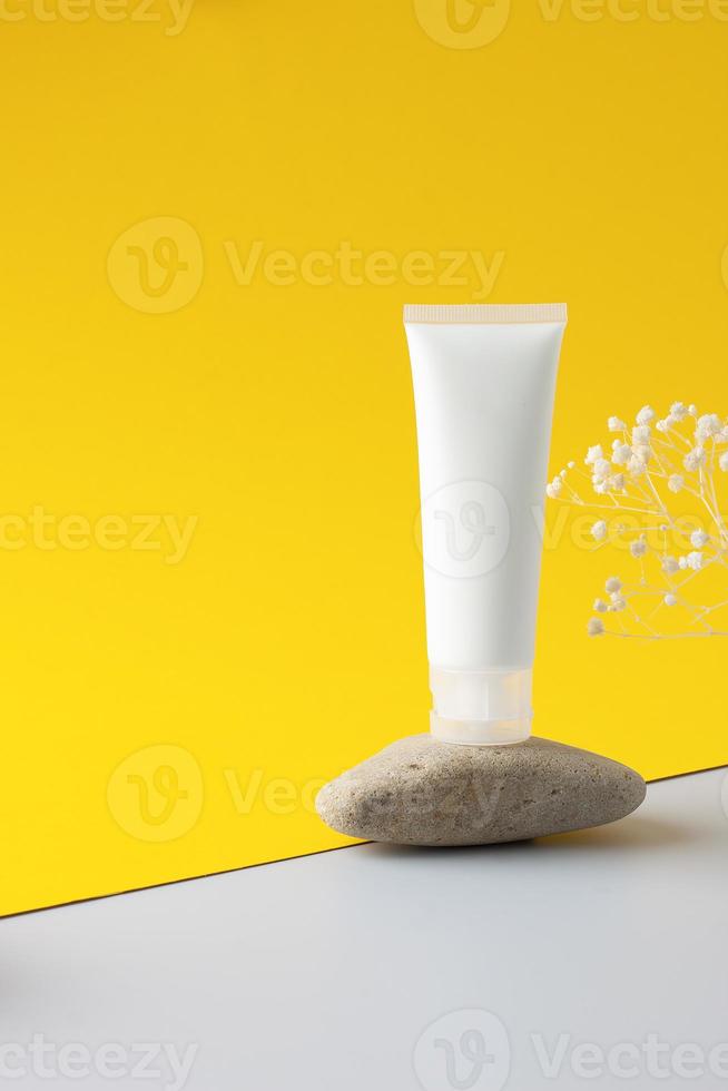 tube blanc de crème cosmétique sur un podium en pierre avec des fleurs sur fond jaune. fermer. photo