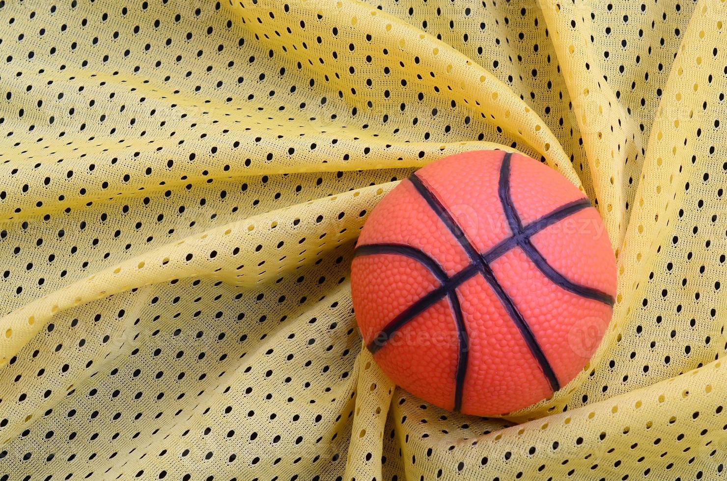 petit basket-ball en caoutchouc orange se trouve sur une texture et un fond de tissu de vêtements de maillot de sport jaune avec de nombreux plis photo