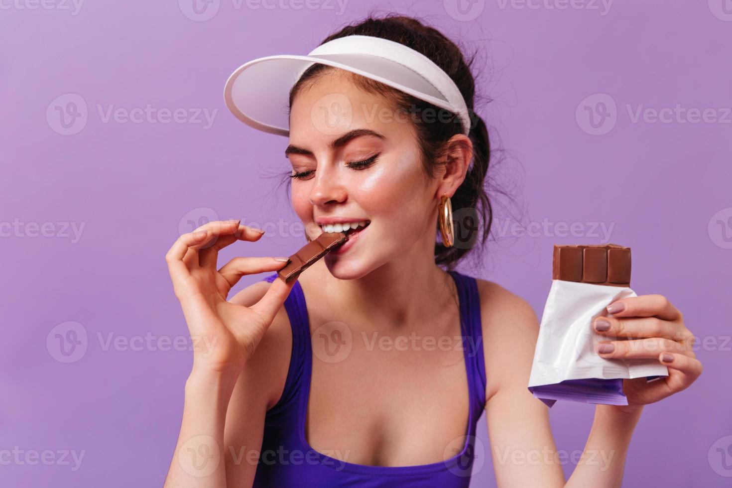 douce fille en casquette et top violet mord avec plaisir du chocolat au lait. brune posant sur le dos violet photo