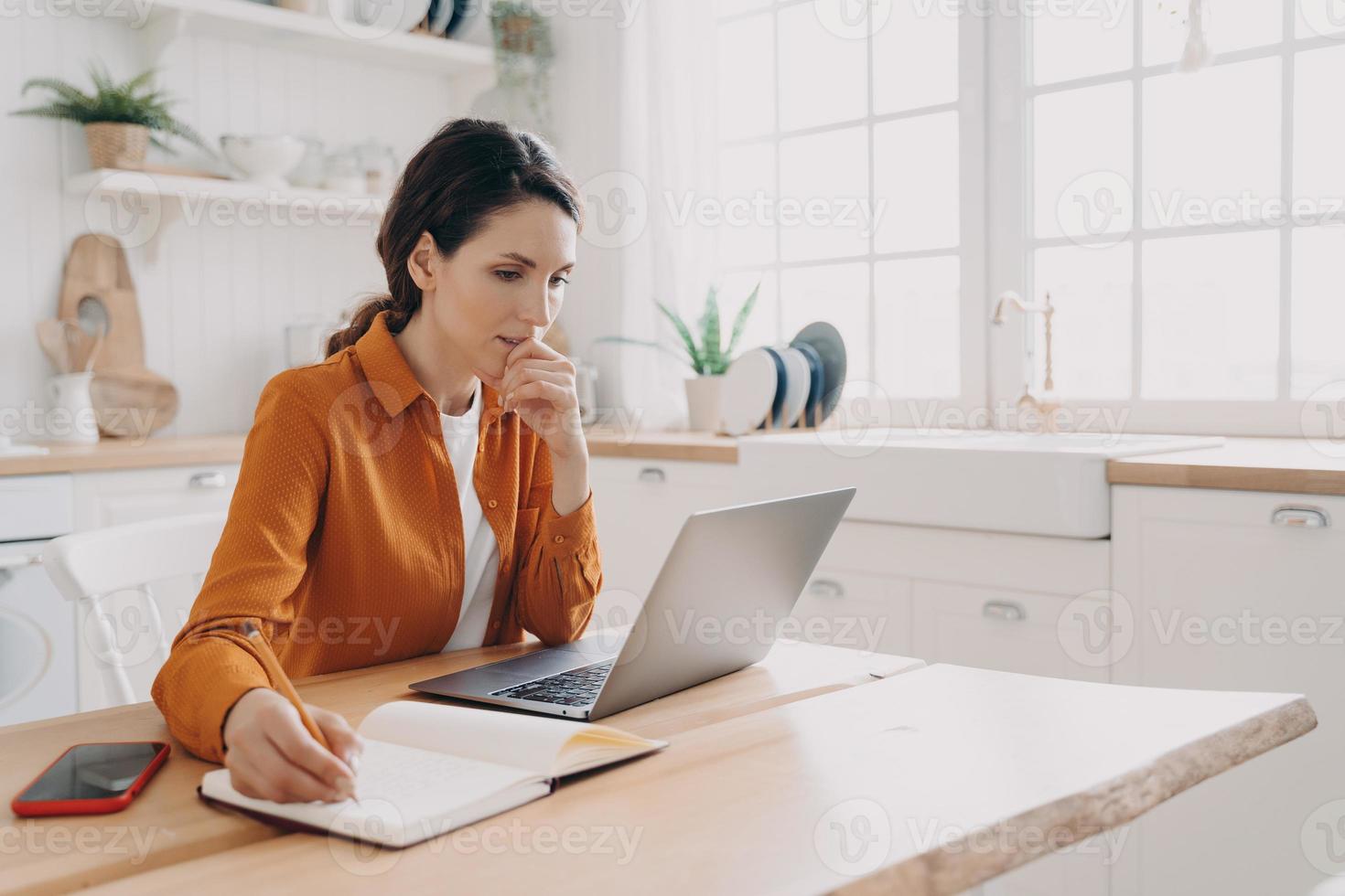 une femme au foyer indépendante travaille sur un ordinateur portable, planifie des achats, prend des notes, est assise dans la cuisine à la maison photo
