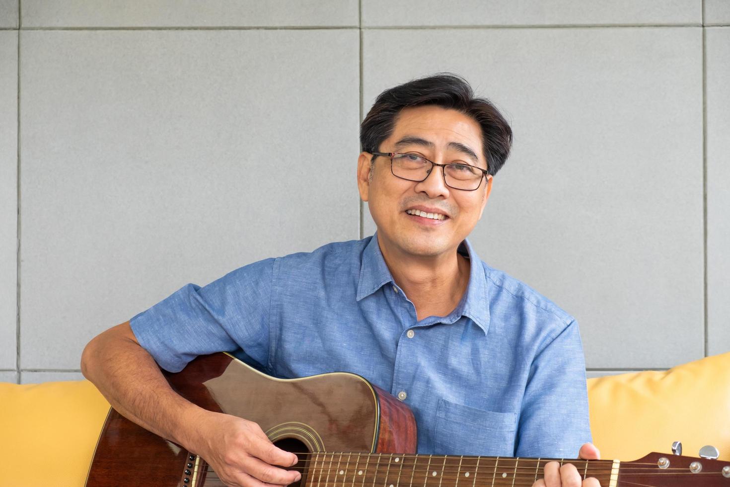 l'homme aîné asiatique aime jouer de la guitare sur le canapé à l'intérieur de la maison de décoration intérieure bien. mode de vie senior actif après la retraite avec bonheur avec une bonne santé mentale. photo