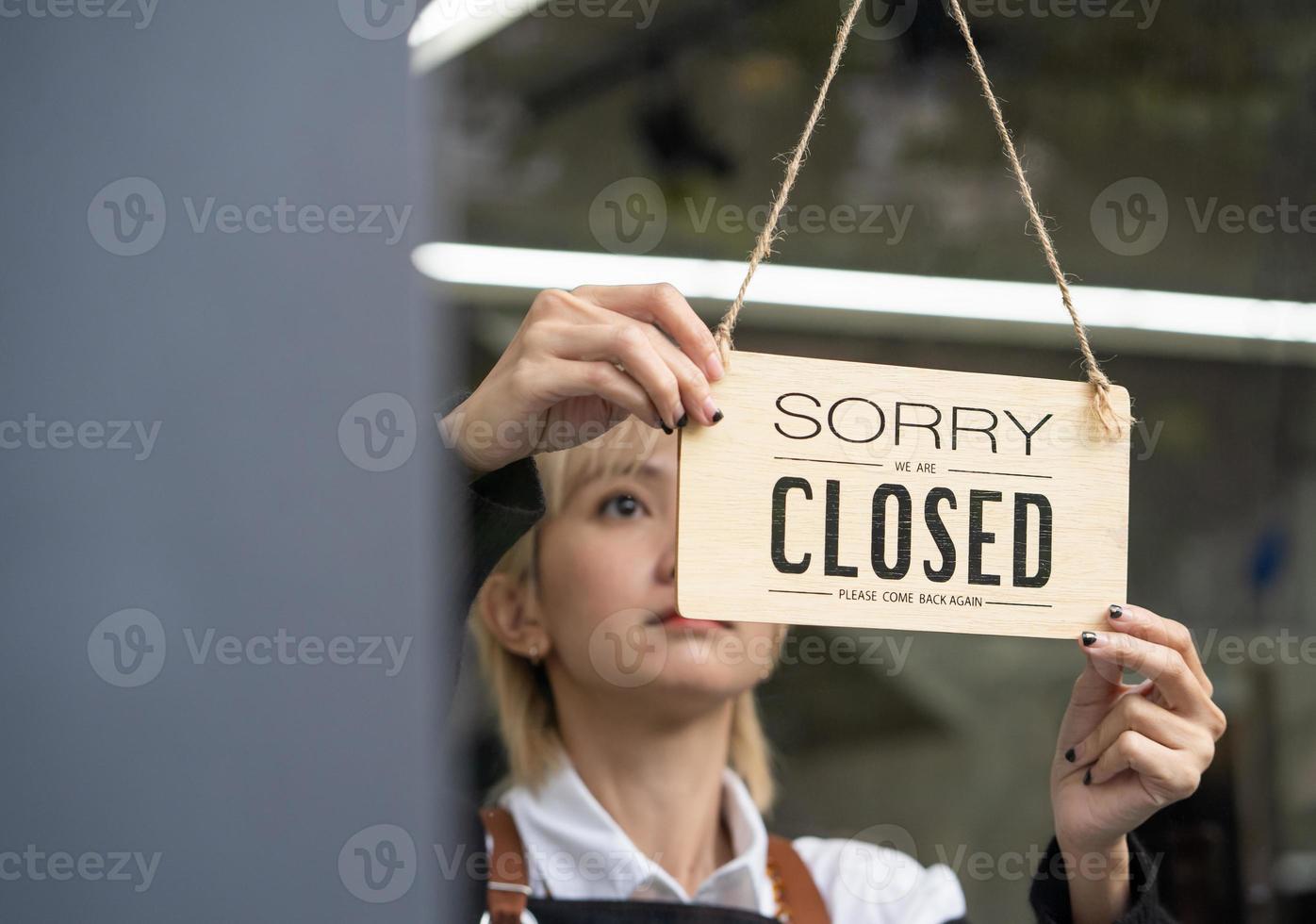 jeune femme propriétaire d'entreprise avec tablier marchant jusqu'à la porte vitrée du magasin pour accrocher un panneau de fermeture à l'entrée avant de fermer le magasin. photo