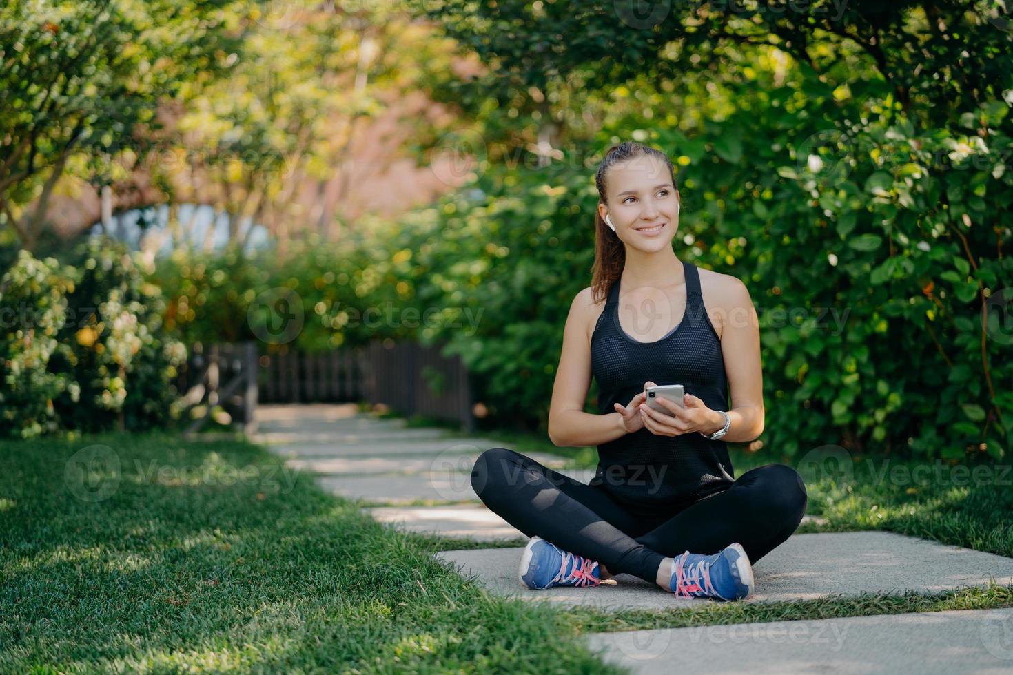 belle sportive est assise les jambes croisées à l'extérieur vêtue de vêtements de sport vérifie le fil d'actualité via un smartphone écoute la musique de la liste de lecture regarde joyeusement loin envoie des messages texte a une formation régulière photo