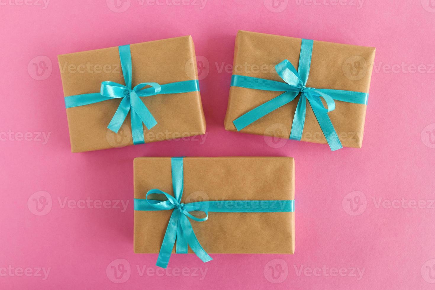 trois coffrets cadeaux enveloppés de papier kraft et ruban bleu sur fond rose, vue de dessus. photo
