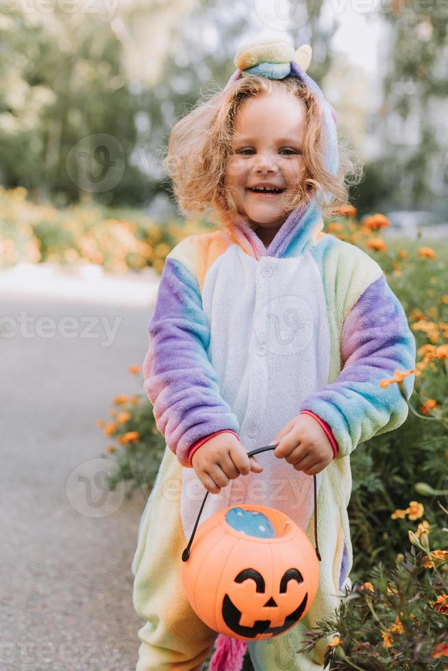 jolie petite fille en costume de licorne arc-en-ciel pour halloween va chercher des bonbons dans un panier de citrouille dans un quartier résidentiel. l'enfant marche à l'extérieur. La charité s'il-vous-plaît. mode de vie. kigurumi photo