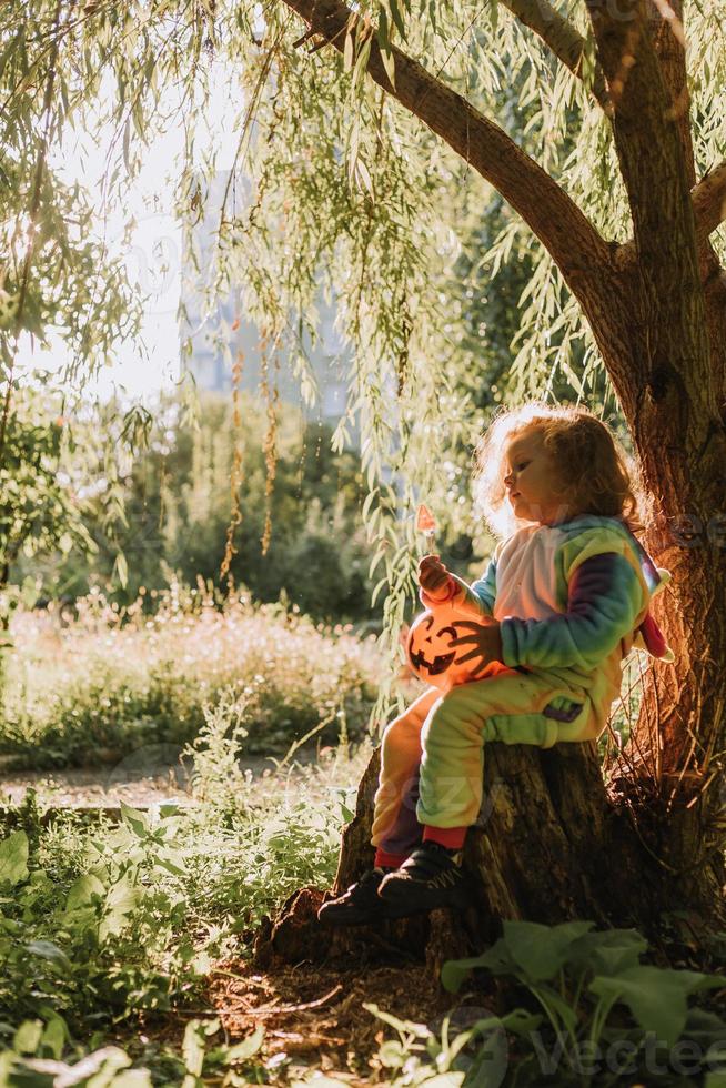 petite fille dans un costume d'halloween de licorne arc-en-ciel et un teckel dans une robe avec un panier de citrouille pour les bonbons sont assis sur une souche au coucher du soleil de la forêt. fabuleuse forêt magique merveilleuse. espace pour le texte photo