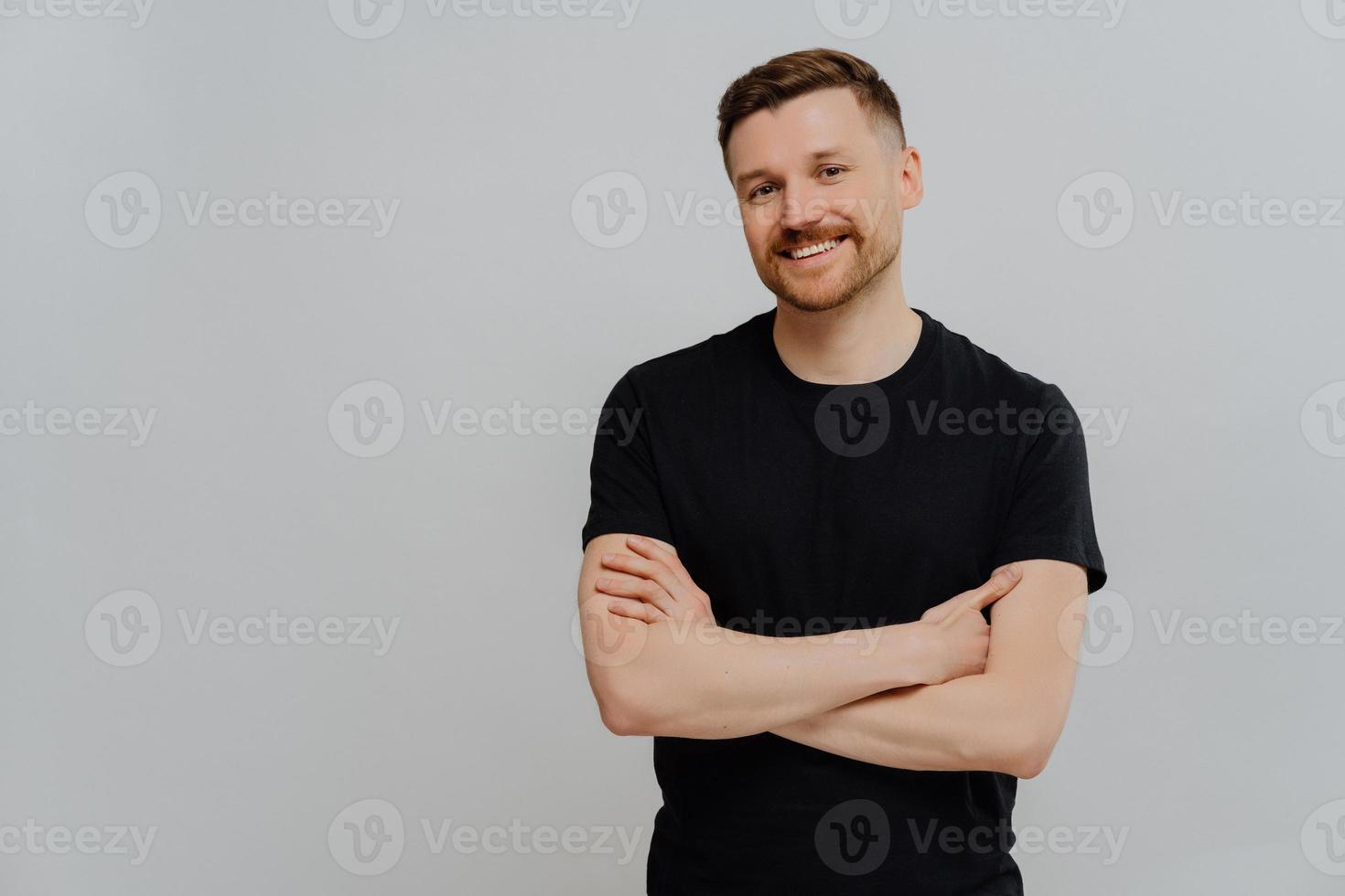 photo horizontale d'un homme satisfait et sûr de lui qui garde les bras croisés, sourit joyeusement habillé avec désinvolture, profite de poses de jour sur fond bleu avec espace vide. beau modèle masculin heureux en studio