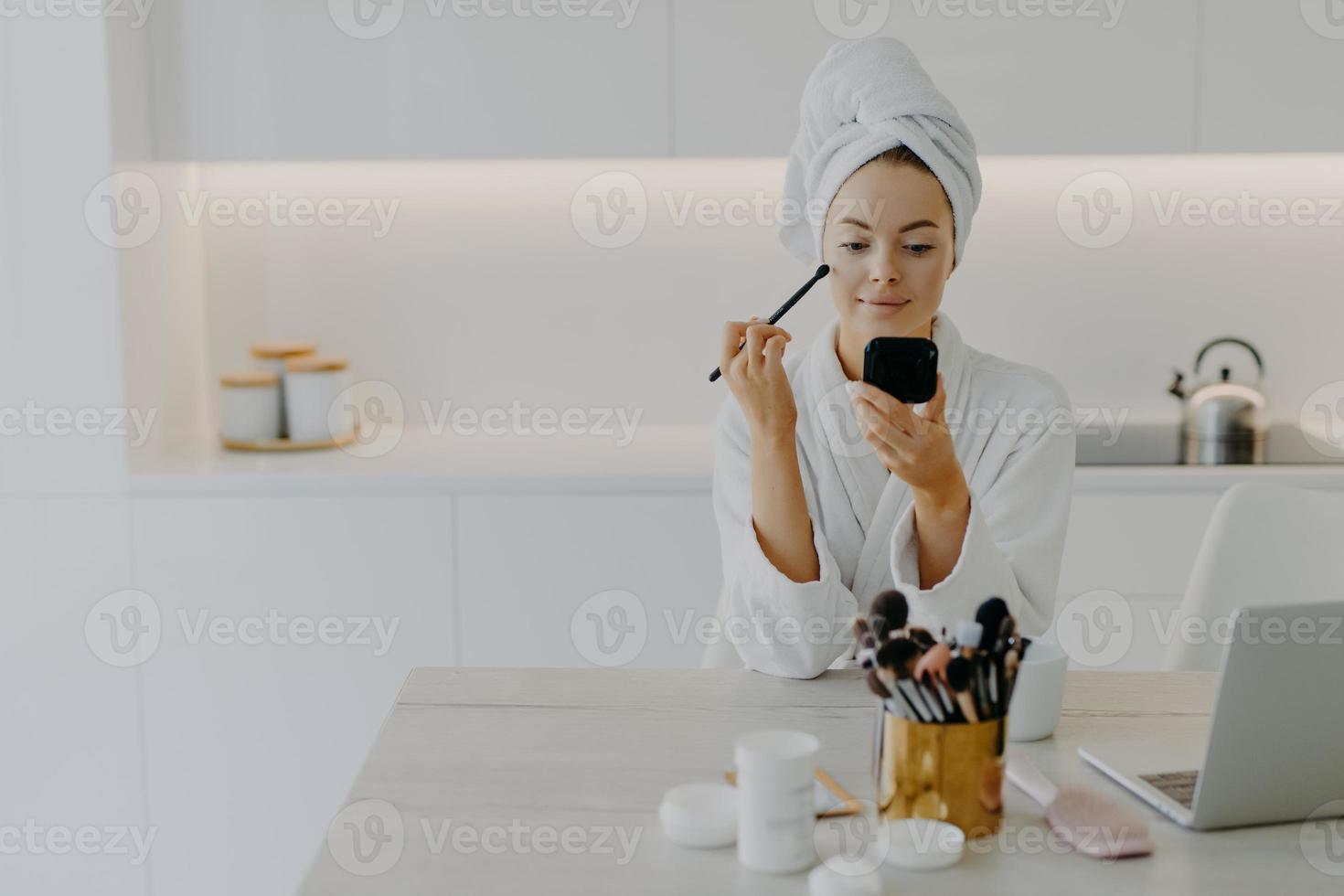 une jeune femme rafraîchie applique de la poudre pour le visage avec un pinceau cosmétique se regarde dans des poses de miroir à la cuisine à la maison est assise au bureau avec des produits de beauté porte une serviette enveloppée et une robe de chambre blanche photo