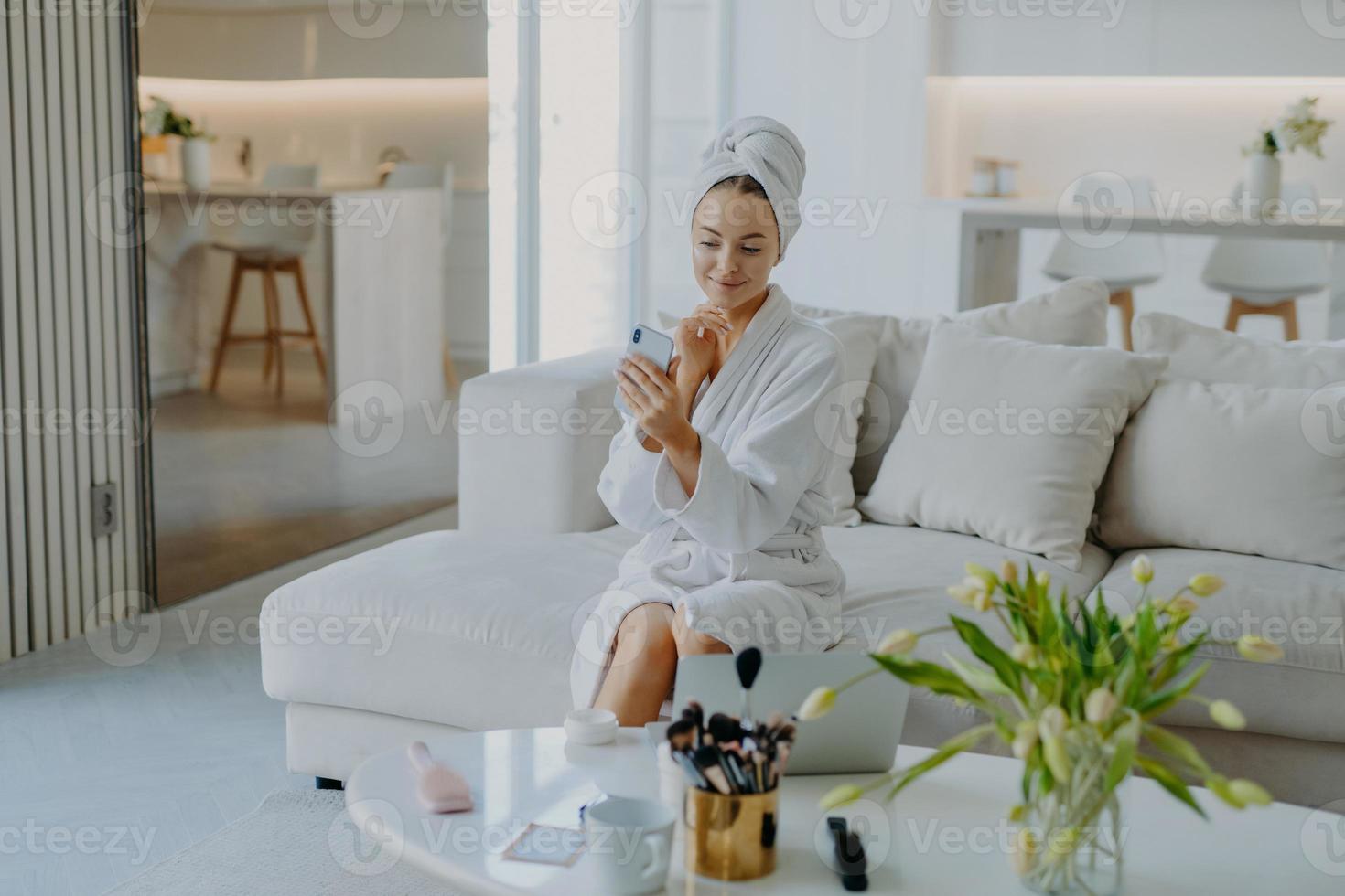 jeune jolie jeune femme regarde un smartphone moderne caméra prend selfie a bien pris soin d'une peau saine applique de la crème porte un peignoir et une serviette pose sur un canapé fait des procédures cosmétiques pour le teint photo