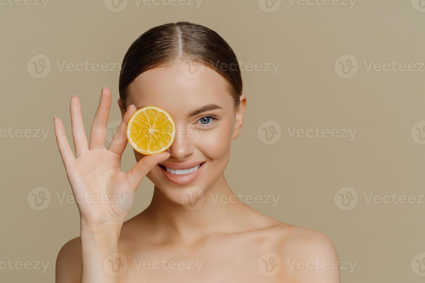 portrait d'une jolie femme brune qui choisit des cosmétiques naturels couvre les yeux avec une tranche de citron frais et juteux sourit à pleines dents propose des produits de beauté bio pour les soins de la peau pose seins nus à l'intérieur. photo