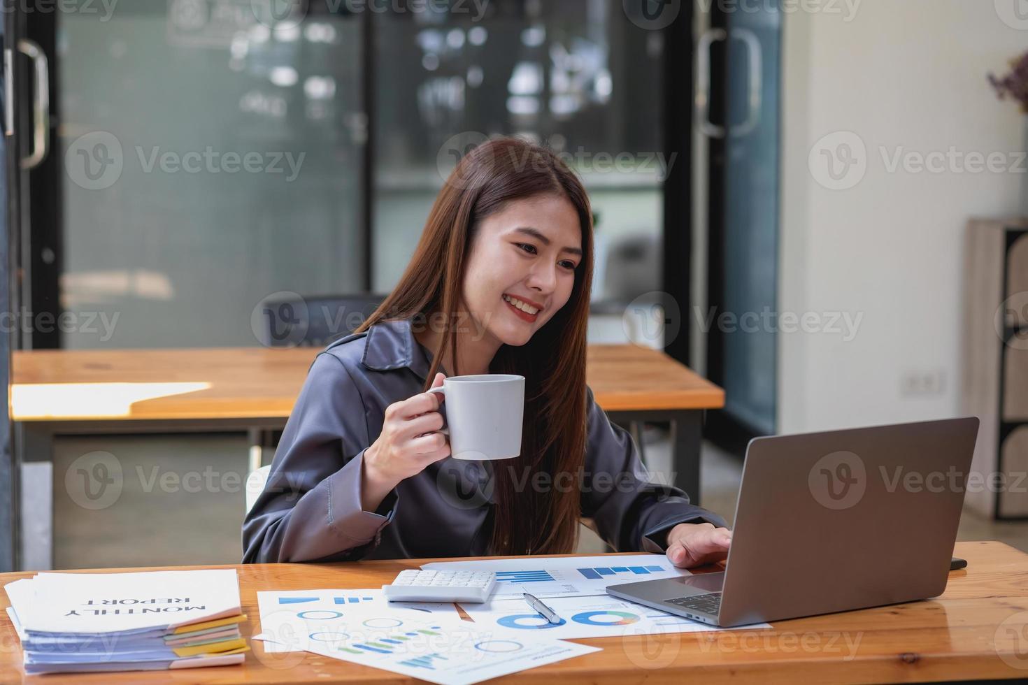 une femme d'affaires tient une tasse de café avant d'analyser les ventes de l'entreprise à l'aide de son ordinateur portable et de documents sur son bureau. photo