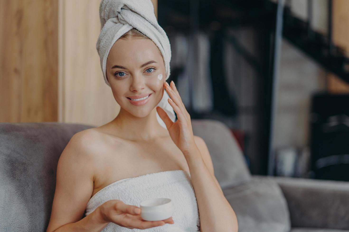 jolie jeune femme à la peau lisse et saine applique de la crème pour le visage, porte une serviette enveloppée sur la tête après avoir pris une douche, pose sur un canapé confortable. procédure d'hygiène, cosmétologie et concept de beauté. photo