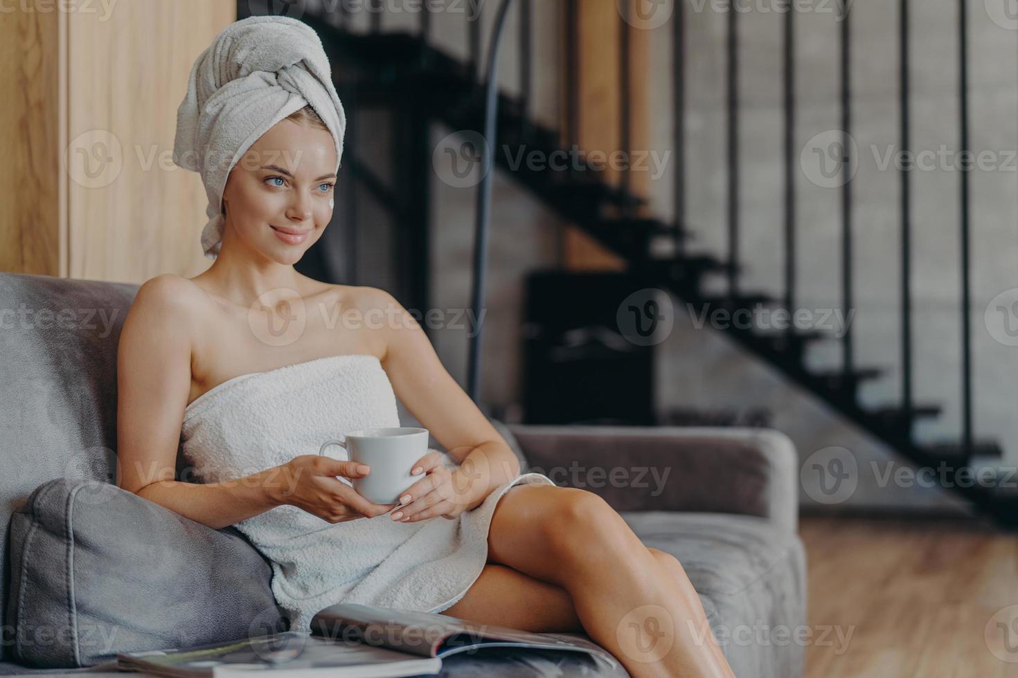 une jeune femme assez détendue regarde attentivement à distance, tient une tasse de boisson chaude, se repose après la douche, enveloppée dans une serviette de bain, s'assoit sur un canapé, lit un magazine, pose contre un intérieur confortable photo