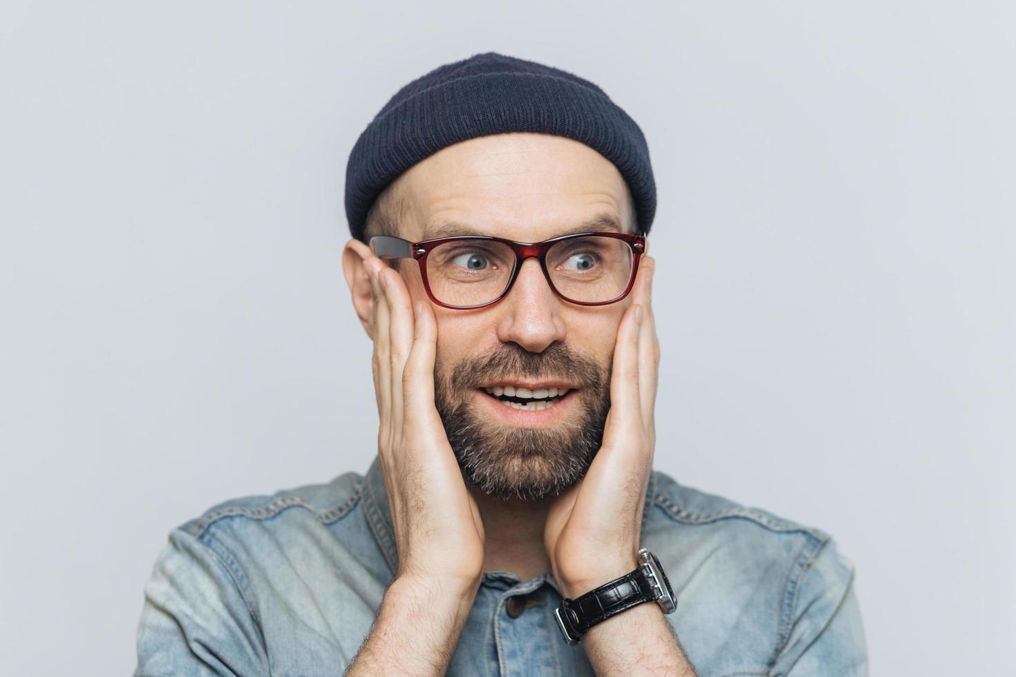 étonné bel homme d'âge moyen regarde à distance dans la perplexité, garde les mains sur les joues, porte des lunettes et un chapeau, isolé sur fond blanc. concept de personnes, d'émotions et d'inattendu photo