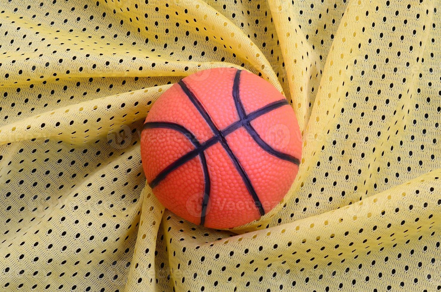 petit basket-ball en caoutchouc orange se trouve sur une texture et un fond de tissu de vêtements de maillot de sport jaune avec de nombreux plis photo