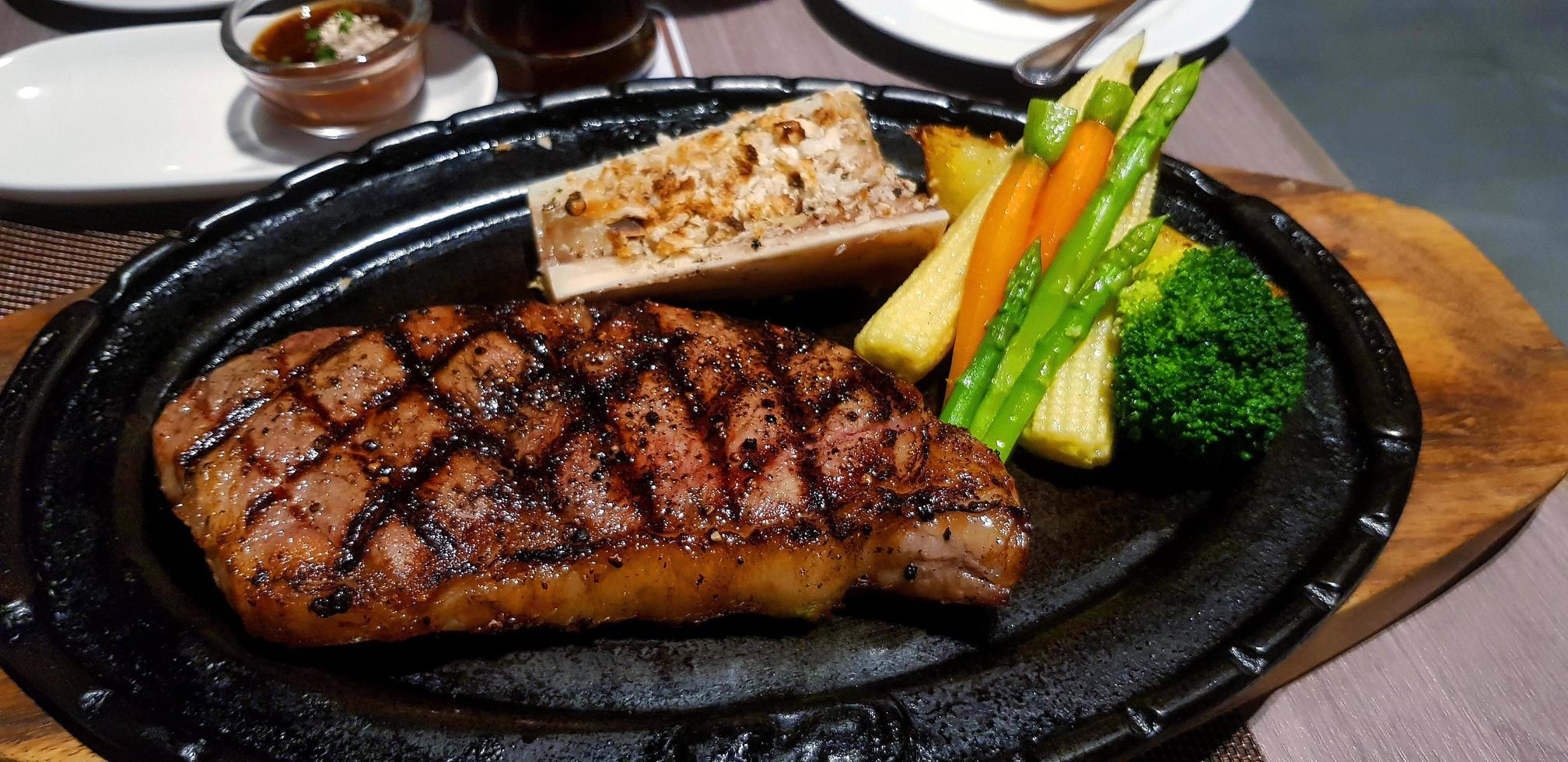 steak de boeuf australien avec carotte, maïs, brocoli et pomme de terre sur une poêle chaude noire. viande grillée ou barbecue avec légumes frais au restaurant. gros repas à l'heure du déjeuner photo