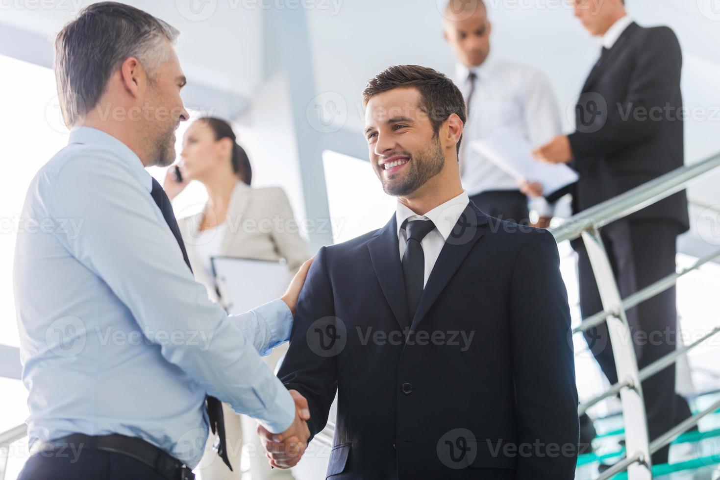 hommes d'affaires se serrant la main. deux hommes d'affaires confiants se serrant la main et souriant tout en se tenant à l'escalier avec des gens en arrière-plan photo