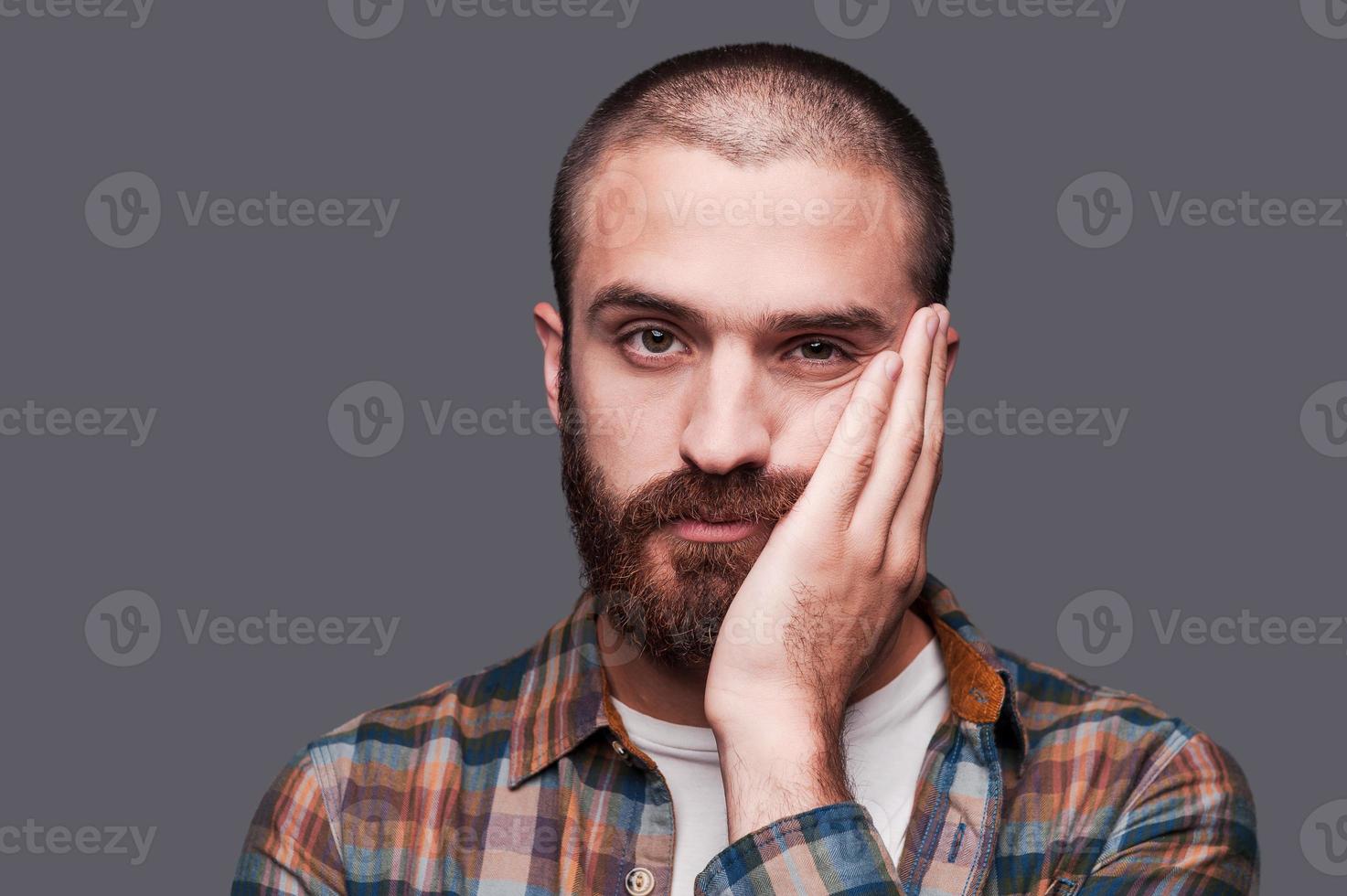 c'est trop ennuyeux. ennuyé jeune homme barbu gardant les bras croisés et exprimant un sourire ironique en se tenant debout sur fond gris photo