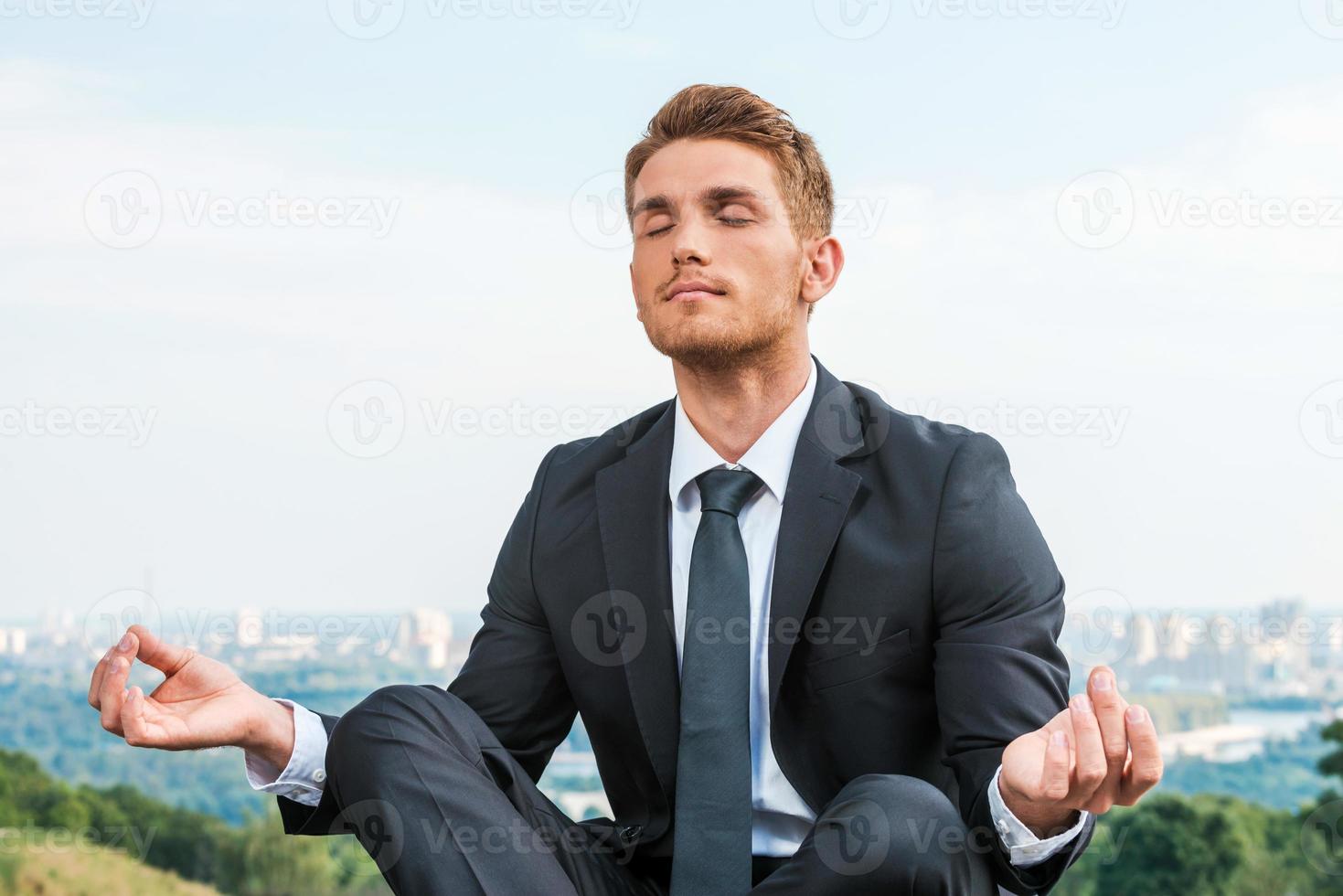 homme d'affaires méditant. jeune homme détendu en tenue de soirée méditant assis en position du lotus avec le paysage urbain en arrière-plan photo