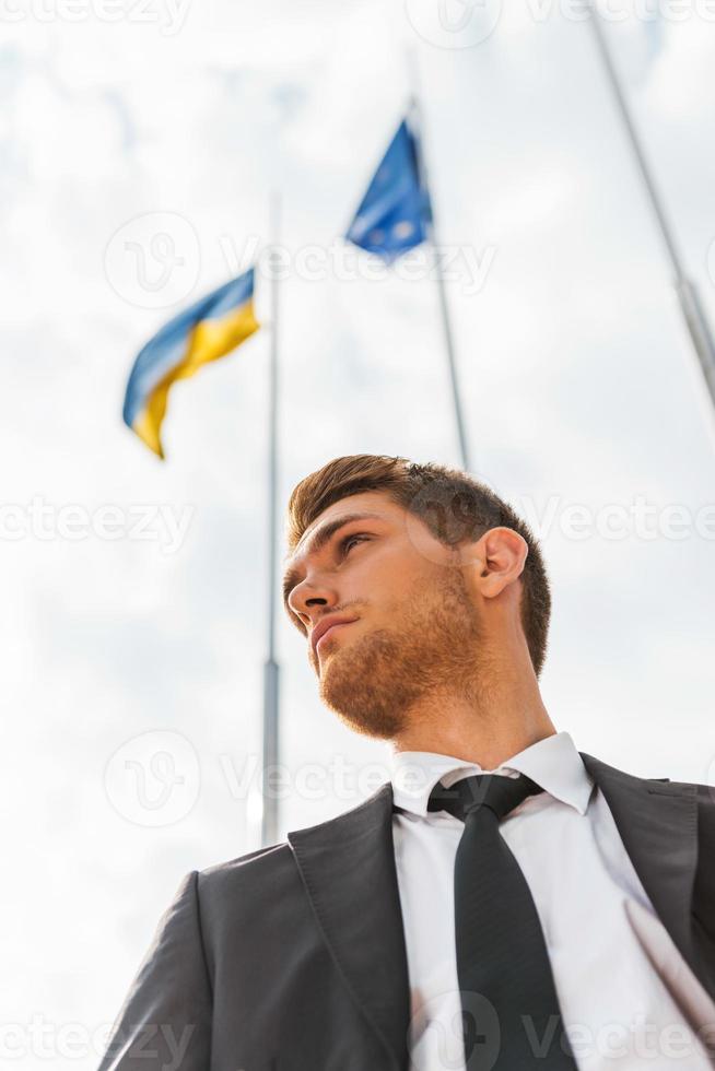 homme d'affaires ukrainien. vue en angle bas d'un jeune homme confiant en tenues de soirée avec des drapeaux ukrainiens et européens en arrière-plan photo