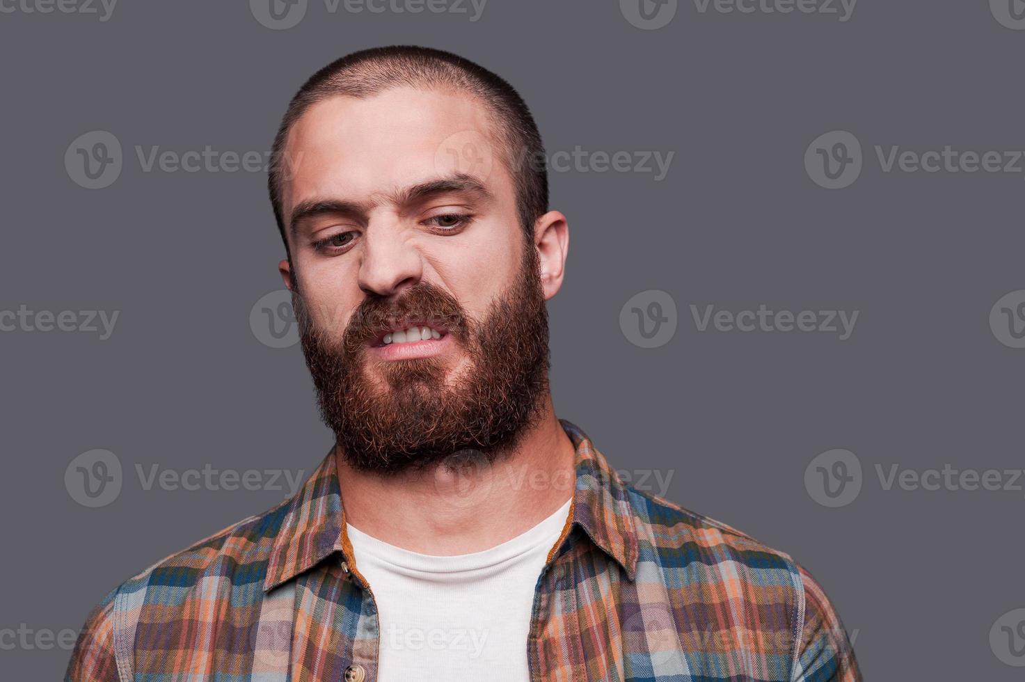 en amis avec ironie. beau jeune homme barbu gardant les bras croisés et exprimant un sourire ironique en se tenant debout sur fond gris photo