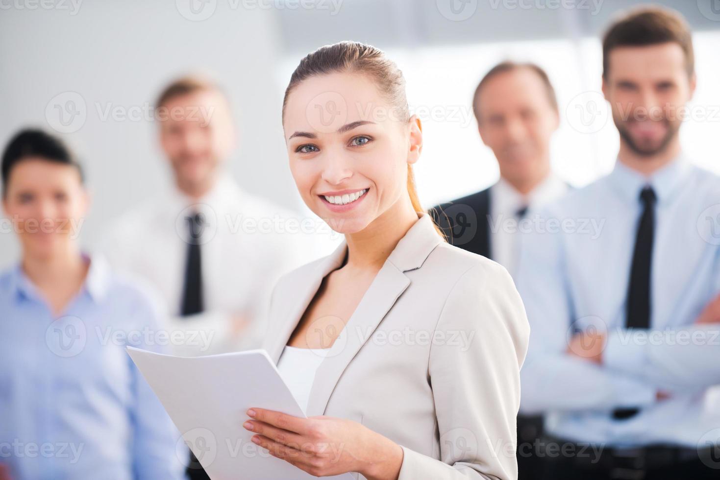 femme d'affaires confiante. séduisante jeune femme d'affaires détenant des documents et souriant tandis que ses collègues se tenant en arrière-plan photo