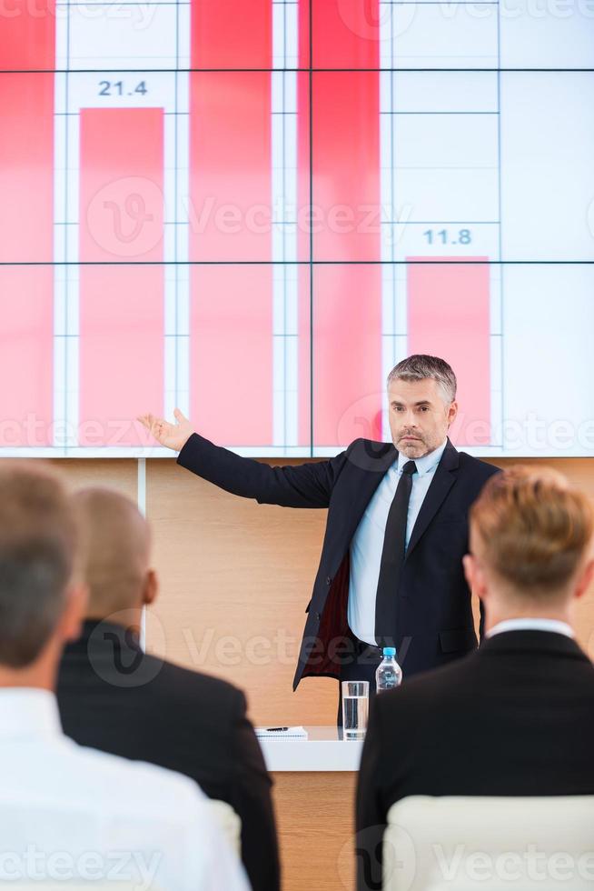 parler des réalisations de l'entreprise. homme mûr confiant en tenues de soirée pointant un écran de projection avec un graphique dessus tout en faisant une présentation dans la salle de conférence avec des personnes au premier plan photo